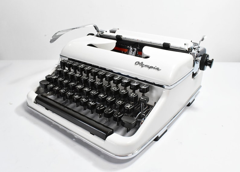 May include: A vintage Olympia typewriter in white with black keys and chrome accents. The typewriter features a classic design with a curved top and the Olympia logo. The keys are arranged in a standard QWERTY layout.