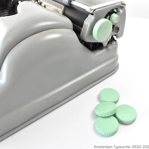 Pode incluir: Uma máquina de escrever cinza com uma tecla verde e cinco tampas de teclas de máquina de escrever verdes sobre uma superfície branca.