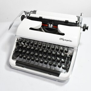 May include: A vintage white Olympia typewriter with black keys and chrome accents. The typewriter has a paper feed and carriage return lever. The word "Olympia" is printed on the front. A classic writing tool.
