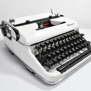 May include: A vintage Olympia typewriter in white with black keys and chrome accents. The typewriter features a classic design with a carriage return lever and a paper feed mechanism. The Olympia logo is visible on the top. A timeless piece for writers and collectors.