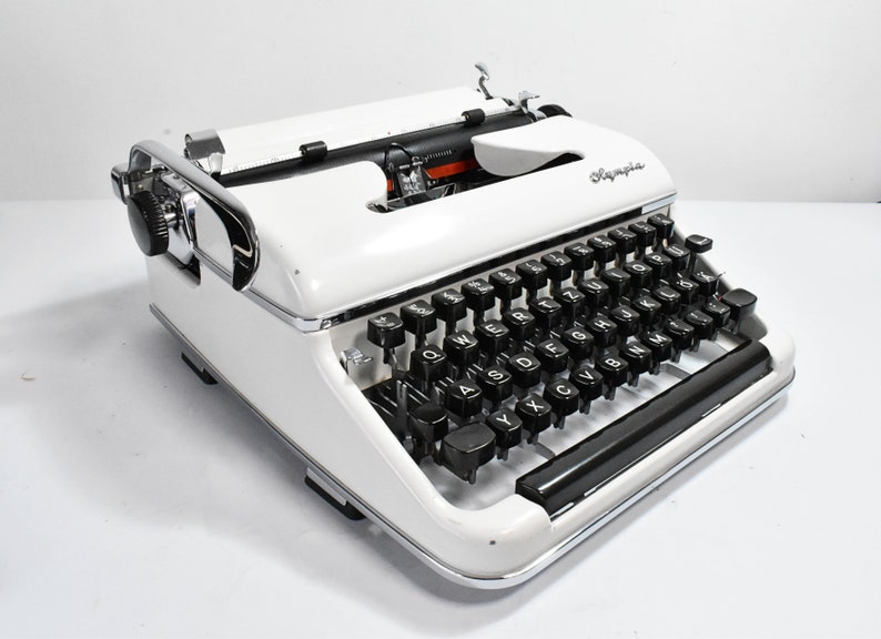 May include: A vintage Olympia typewriter in white with black keys and chrome accents. The typewriter features a classic design with a carriage return lever and a paper feed mechanism. The Olympia logo is visible on the top. A timeless piece for writers and collectors.