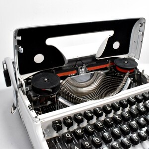 May include: A vintage white typewriter with black keys and a silver carriage. The top cover is open, revealing the internal mechanisms, including the ribbon spools and typebars. The keys are arranged in a QWERTY layout.