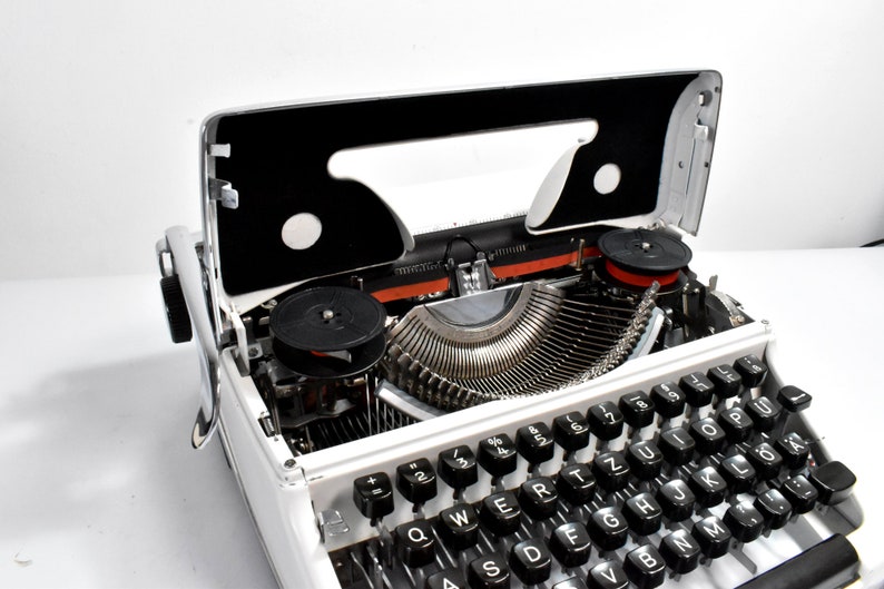 May include: A vintage white typewriter with black keys and a silver carriage. The top cover is open, revealing the internal mechanisms, including the ribbon spools and typebars. The keys are arranged in a QWERTY layout.