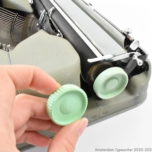 Pode incluir: Close-up de uma maçaneta de plástico verde em uma máquina de escrever vintage. A maçaneta está sendo segurada por uma mão.