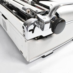 May include: Close-up of a vintage white typewriter with chrome accents. The typewriter features a black knob, a curved chrome handle, and a detailed mechanical design. The image highlights the machine's classic aesthetic.