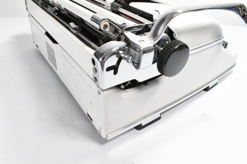 May include: Close-up of a vintage white typewriter with chrome accents. The typewriter features a black knob, a curved chrome handle, and a detailed mechanical design. The image highlights the machine's classic aesthetic.