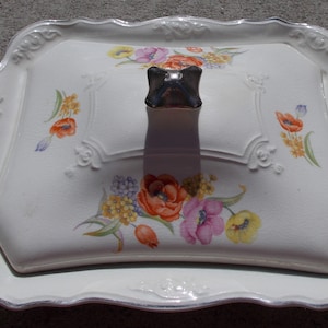 Può includere: Piatto da portata in ceramica bianca con bordo ondulato e motivo floreale. Il piatto ha un coperchio con un pomo argentato.