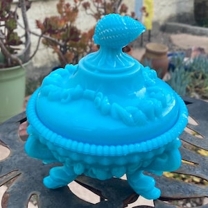 May include: A bright turquoise blue glass candy dish featuring a lid topped with a seashell finial. The bowl and lid are decorated with embossed shell motifs and a beaded rim, supported by three ornate feet in a matching vibrant blue glass.