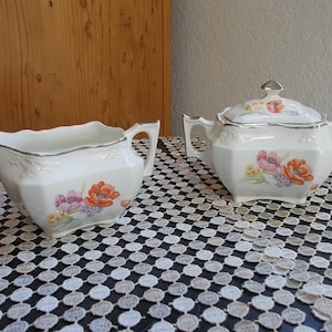 May include: A white porcelain creamer and sugar bowl set with floral designs. The creamer has a silver rim and the sugar bowl has a silver lid.
