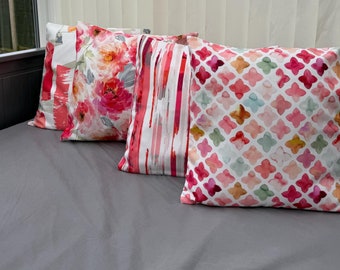 May include: Three decorative throw pillows with floral and geometric patterns in shades of pink, red, and white. The pillows are arranged on a gray couch.