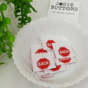 May include: A white bowl filled with ten white woven labels featuring red stop signs that say "BACK" in white lettering. The labels are designed for clothing and are sold by Josie Buttons.
