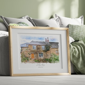 May include: A framed watercolor print of a stone cottage with a brown door and climbing vines. The artwork is set in a light wood frame and features the text "Holly Cottage" and "February 2024". The print is displayed on a bed with pillows.