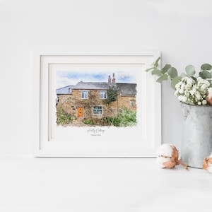 May include: A framed watercolor print of a stone cottage with an orange door and climbing plants. The print is in a white frame and is labeled "Holly Cottage" with the date "February 2004". A galvanized metal bucket with white flowers is to the right.