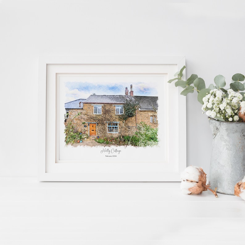 May include: A framed watercolor print of a stone cottage with an orange door and climbing plants. The print is in a white frame and is labeled "Holly Cottage" with the date "February 2004". A galvanized metal bucket with white flowers is to the right.