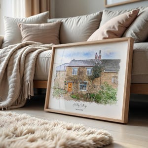 May include: A framed watercolor print of a stone cottage with a wooden frame. The cottage has a brown door, multiple windows, and is covered in vines. The print is labeled "Holly Cottage" and "February 2024". The frame is resting on a light-colored floor.