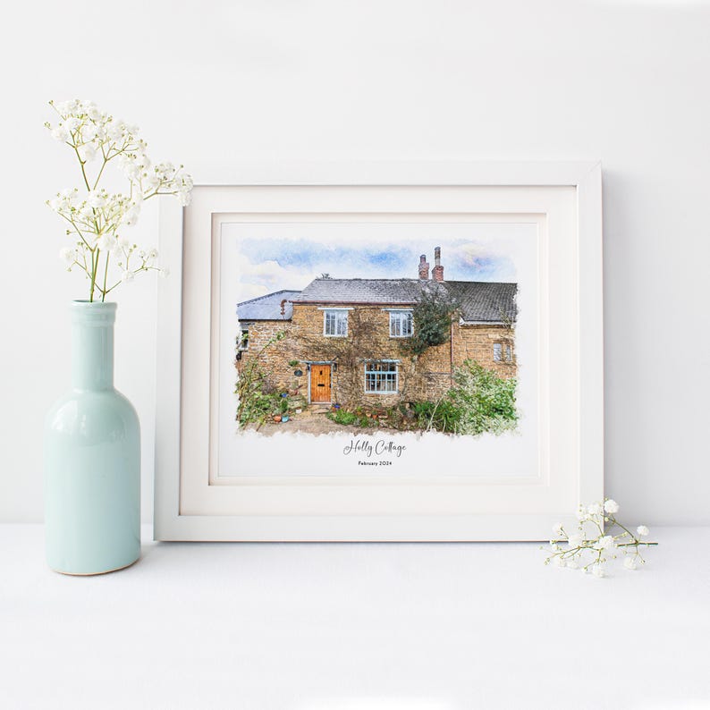 May include: A framed watercolor print of a stone cottage with a yellow door and climbing vines. The print is in a white frame and has the text "Holly Cottage" and "February 2024" at the bottom. A light blue vase with white flowers is on the left.