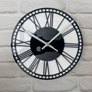 May include: A circular wall clock featuring a black frame and Roman numerals. The clock face is white with a black central disc. The clock hands are silver. The clock is mounted on a white brick wall.