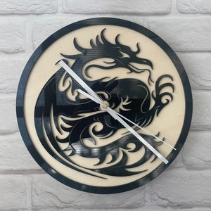 May include: A black and white clock featuring a dragon design. The dragon is curled up with its wings outstretched. The clock face is white with black hands.