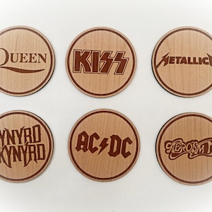 May include: Set of six wooden coasters featuring iconic rock band logos: Queen, Kiss, Metallica, Lynyrd Skynyrd, AC/DC, and Aerosmith. Each coaster has a dark brown border and text, with a natural wood grain background.
