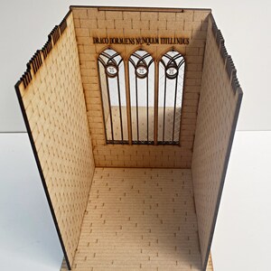 Op de afbeelding: Een houten model van een kamer met een bakstenen muur en een groot raam. Het raam heeft drie gebogen secties met de tekst "DRACO DORMIENS NUNQUAM TITTILLANDUS" erboven.
