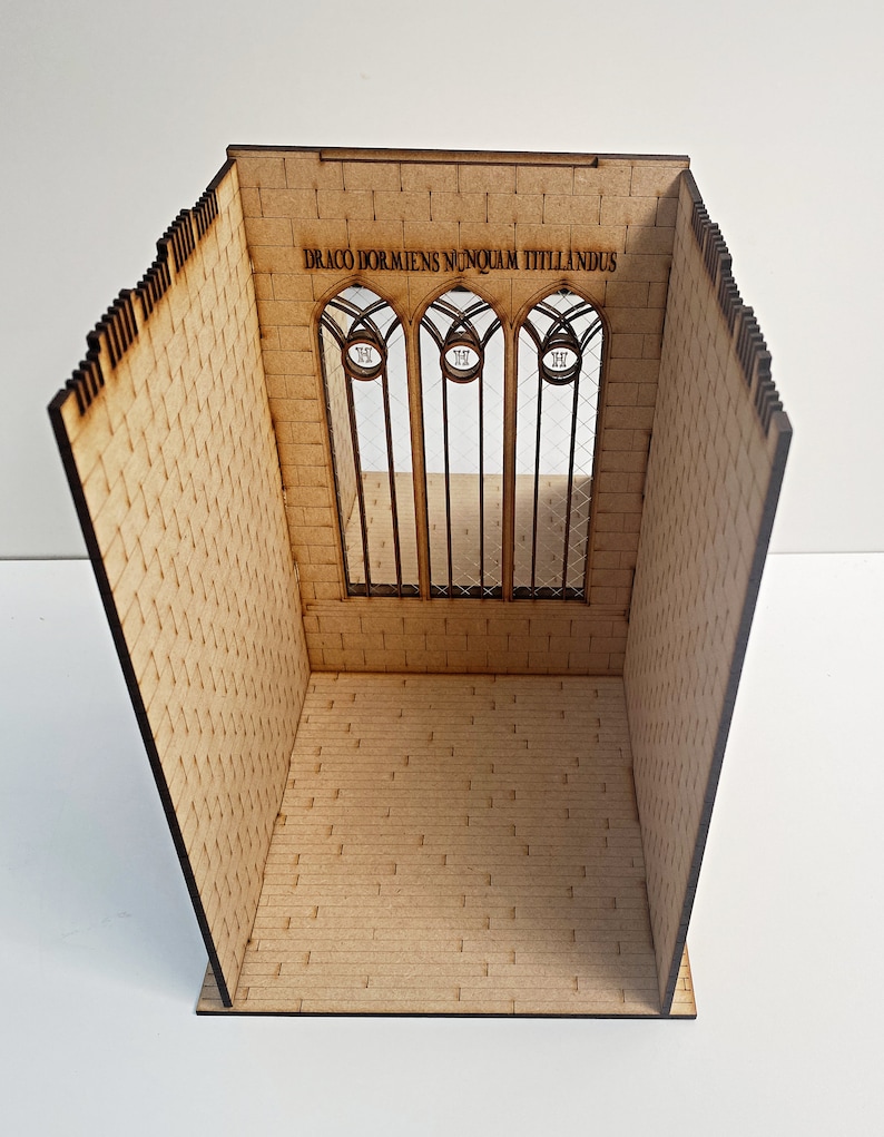 Op de afbeelding: Een houten model van een kamer met een bakstenen muur en een groot raam. Het raam heeft drie gebogen secties met de tekst "DRACO DORMIENS NUNQUAM TITTILLANDUS" erboven.