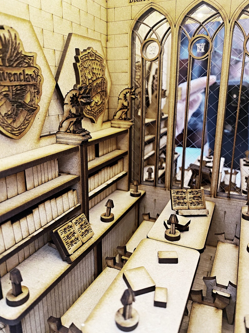 Op de afbeelding: Een houten model van een bibliotheek van Zweinstein met vier huiswapens, boekenkasten, bureaus en lampen. De wapens zijn voor Griffoendor, Huffelpuf, Zwadderich en Ravenklauw.