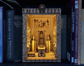 Puede incluir: Un diorama en miniatura de un templo egipcio de madera con iluminación interior. La estructura detallada presenta jeroglíficos, estatuas y un símbolo ankh central. El diorama se exhibe en una estantería con varios libros.