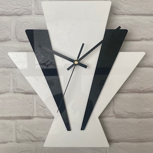 Op de afbeelding: Een geometrische wandklok in zwart en wit met gouden wijzers. De klok heeft de vorm van een waaier met overlappende driehoeken.