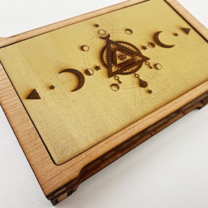 May include: A rectangular wooden box featuring a light-colored wood frame and a central panel with a laser-engraved design. The artwork displays an eye within a triangle, surrounded by moon phases, stars, and geometric lines in dark brown tones.