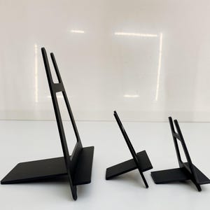 May include: Three black metal display stands of varying sizes are arranged on a white surface. The stands have a rectangular base and a vertical support with a cutout design. The largest stand is approximately 25 cm tall.