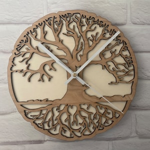 Op de afbeelding: Een ronde houten wandklok met een levensboomontwerp. De klok heeft een lichtbruin frame met een uitgesneden boom silhouet en witte wijzers. De klok is gemonteerd op een witte bakstenen muur.