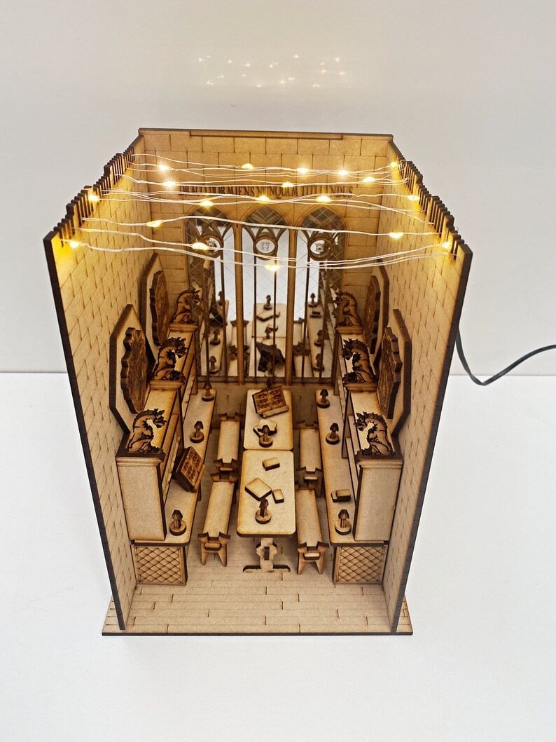 Op de afbeelding: Een houten model van een klaslokaal van Zweinstein met een lange tafel en stoelen, verlicht door lichtslingers. De muren zijn versierd met steenwerk en de ramen zijn gebogen. De tekst "DRACO DORMIENS NUNQUAM TItillANDUS" is zichtbaar op de muur.