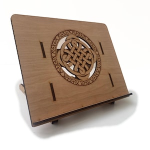 May include: A wooden book stand with a Celtic knot design. The stand is made of light-coloured wood and features a laser-cut knot design in the centre. It has a sloped surface to hold books and a small ledge at the bottom to keep pages in place.