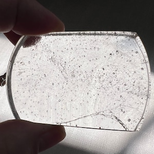May include: A clear glass pendant with a rounded shape and a textured surface. The glass has small air bubbles trapped inside.