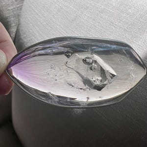 May include: A clear, oval-shaped crystal with a faceted interior. The crystal has a purple hue on one end and a clear, reflective surface. The object is held against a light gray background.