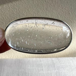 May include: A clear oval-shaped crystal with a silver rim. The crystal has a textured surface with small bubbles throughout.