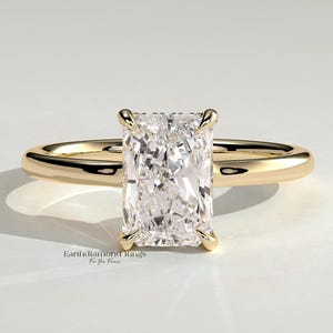 May include: A gold ring featuring a large, rectangular radiant-cut center stone held by four prongs. The band is smooth, polished yellow gold. The text Earthdiamond Rings For You Forever is printed in small black letters below the ring.