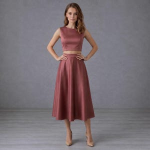 May include: A two-piece set consisting of a sleeveless, round-neck crop top and a matching high-waisted, A-line midi skirt. The garment is made of a solid, dusty rose-coloured fabric with a smooth, satin-like finish and a subtle sheen.