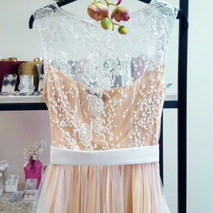 Puede incluir: Un vestido de tul color melocot&oacute;n con una superposici&oacute;n de encaje blanco y una faja de sat&eacute;n blanco. El vestido es sin mangas y tiene un escote redondo.