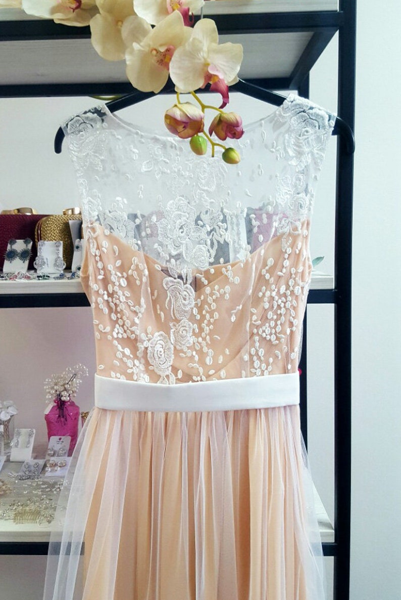 Puede incluir: Un vestido de tul color melocot&oacute;n con una superposici&oacute;n de encaje blanco y una faja de sat&eacute;n blanco. El vestido es sin mangas y tiene un escote redondo.