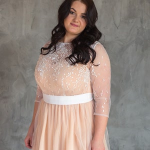 May include: A woman wearing a peach-coloured tulle dress with white lace detailing and a white sash. She has long dark hair and is looking at the camera.