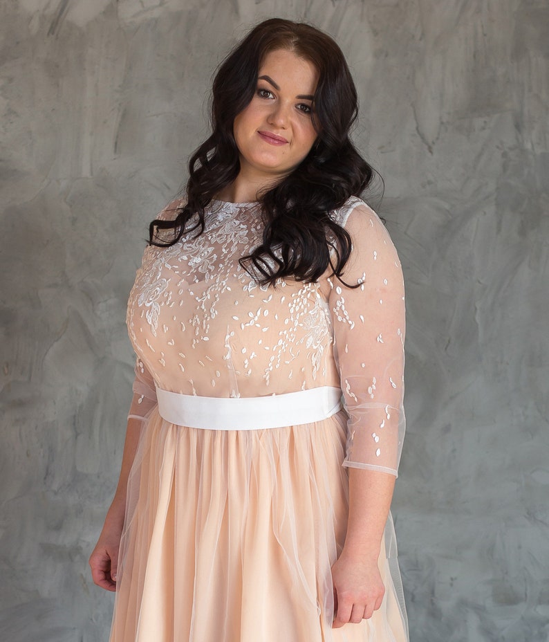 May include: A woman wearing a peach-coloured tulle dress with white lace detailing and a white sash. She has long dark hair and is looking at the camera.