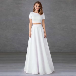 Puede incluir: Un conjunto formal blanco de dos piezas. La parte superior es un top corto de manga corta con cuello redondo. La falda es un diseño largo hasta el suelo, de línea A. El conjunto está hecho de una tela lisa y opaca. El fondo es gris liso.
