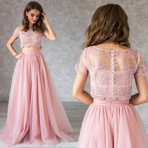 Puede incluir: Un conjunto de dos piezas en color rosa, compuesto por una falda de tul y una blusa de encaje. La blusa tiene mangas cortas y un diseño de encaje. La falda es amplia y fluida.