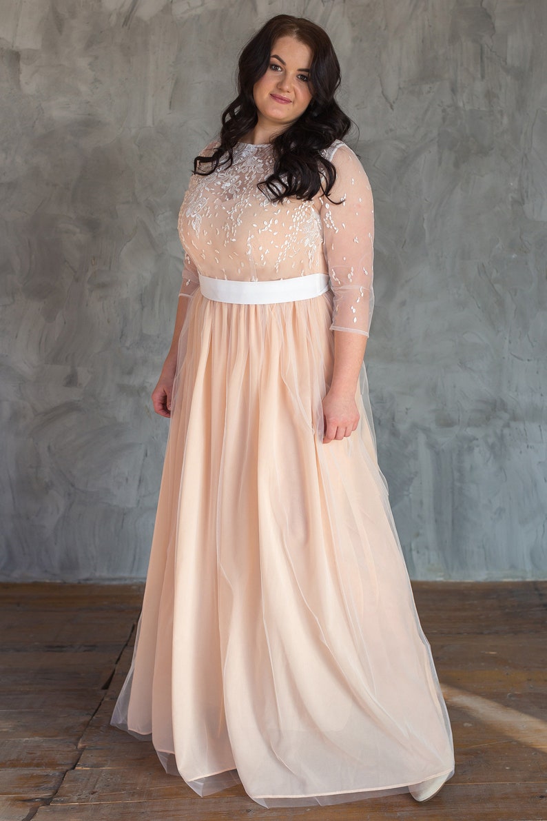 May include: A woman wearing a peach tulle gown with a white lace bodice and a white satin sash. The gown has a long skirt and is floor-length.