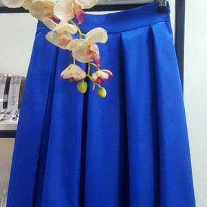 May include: A vibrant blue skirt with pleats, hanging on a hanger. The skirt is made of a smooth fabric and is displayed with a white and pink orchid arrangement. The skirt is a solid color and appears to be knee-length.