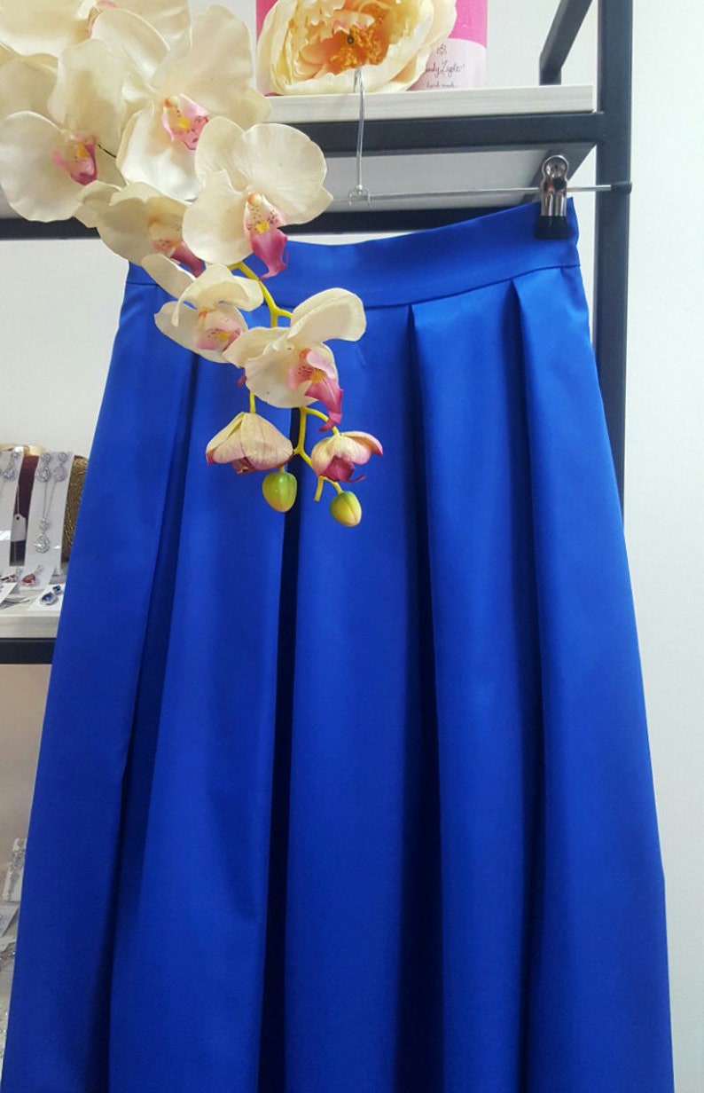 May include: A vibrant blue skirt with pleats, hanging on a hanger. The skirt is made of a smooth fabric and is displayed with a white and pink orchid arrangement. The skirt is a solid color and appears to be knee-length.