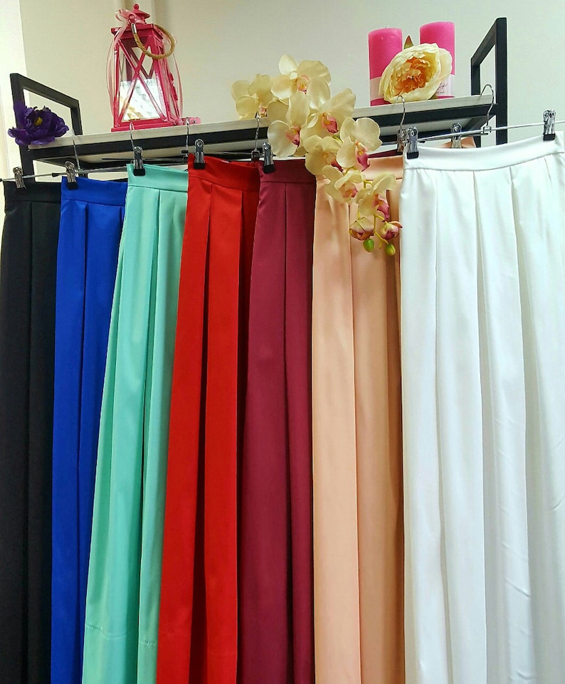 May include: A display of colorful, pleated skirts hanging on a rack. The skirts are in various colors, including black, blue, teal, red, burgundy, peach, and white. The skirts are made of a smooth, flowing fabric.