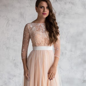May include: A woman wearing a blush-coloured tulle dress with a white lace bodice and a white satin sash. The dress has long sleeves and a flowing skirt.