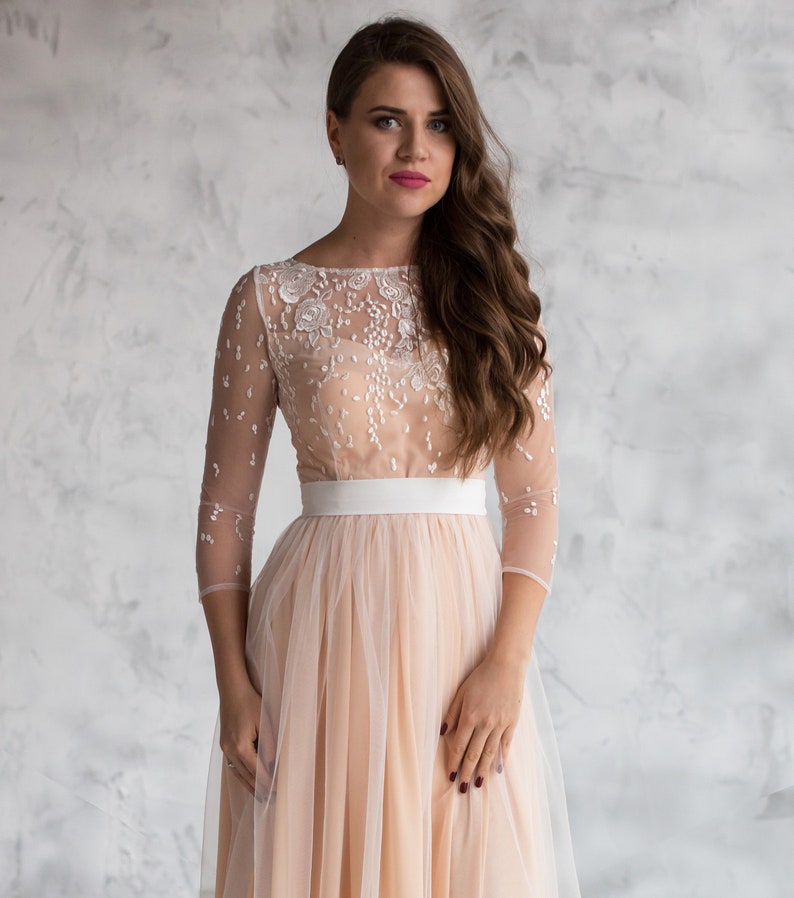May include: A woman wearing a blush-coloured tulle dress with a white lace bodice and a white satin sash. The dress has long sleeves and a flowing skirt.